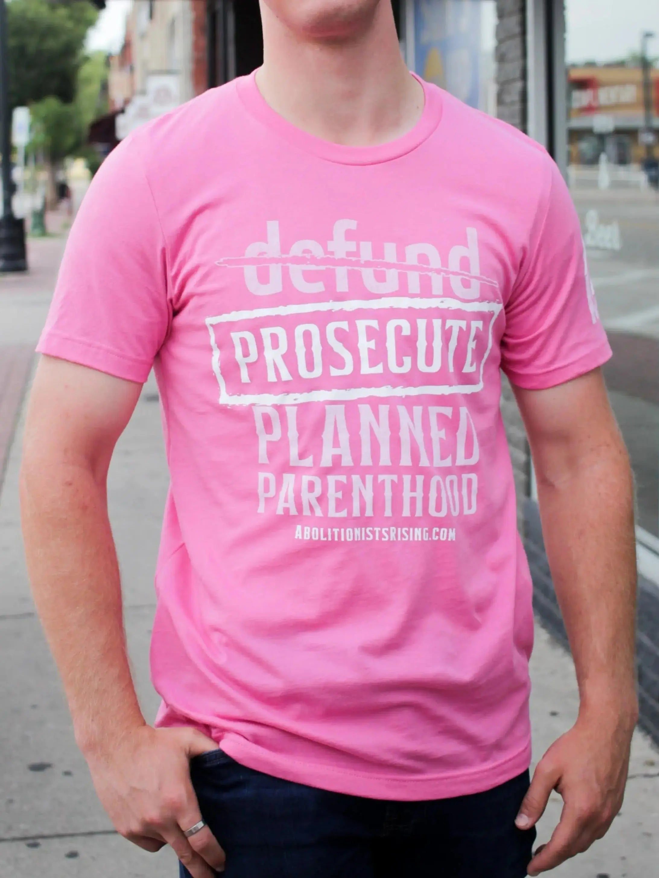 Person wearing a pink t-shirt with defund prosecute planned parenthood text on a city street.