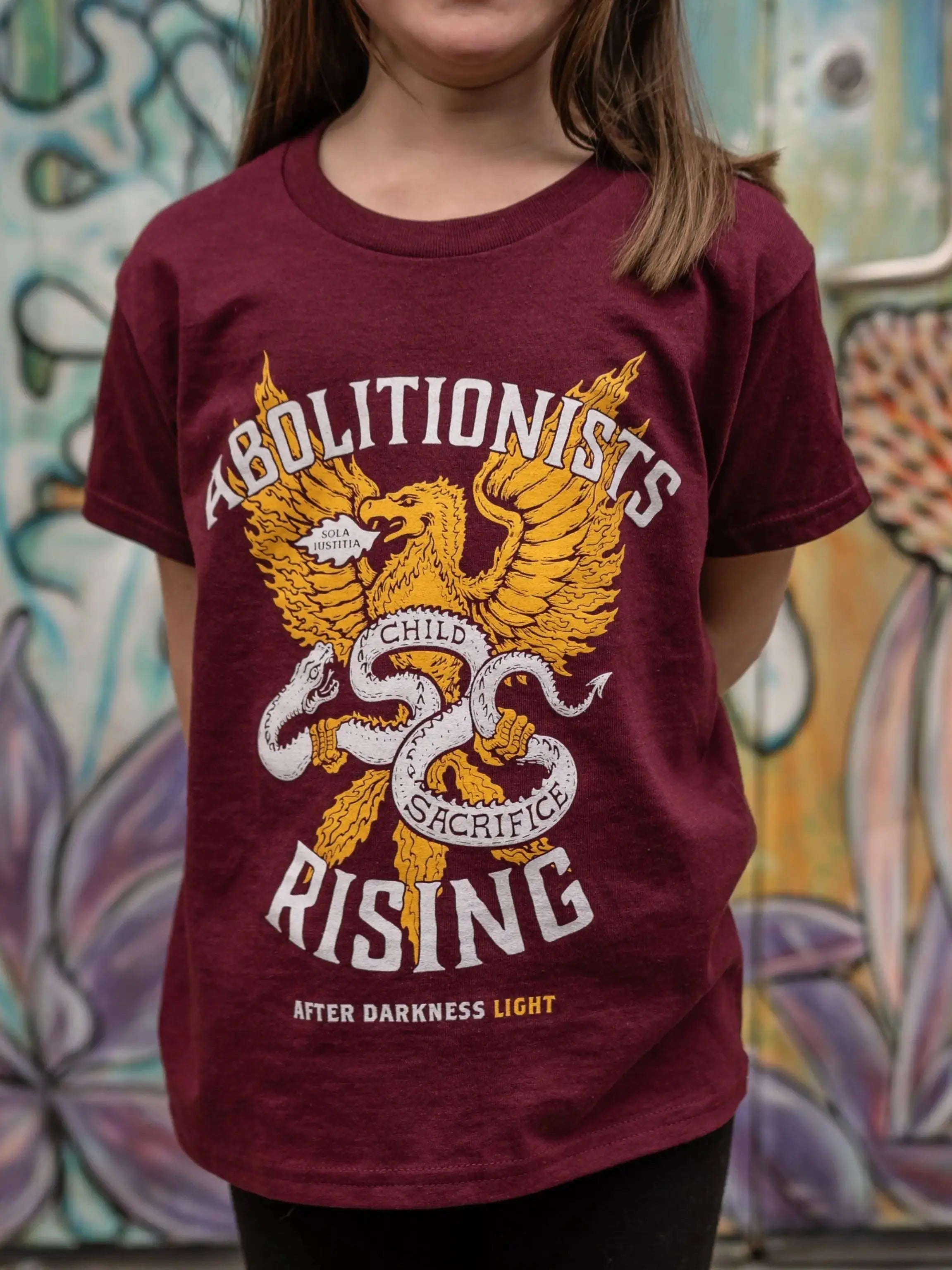 Child wearing a maroon t-shirt with graphic Abolitionists Rising design in front of a colorful mural.